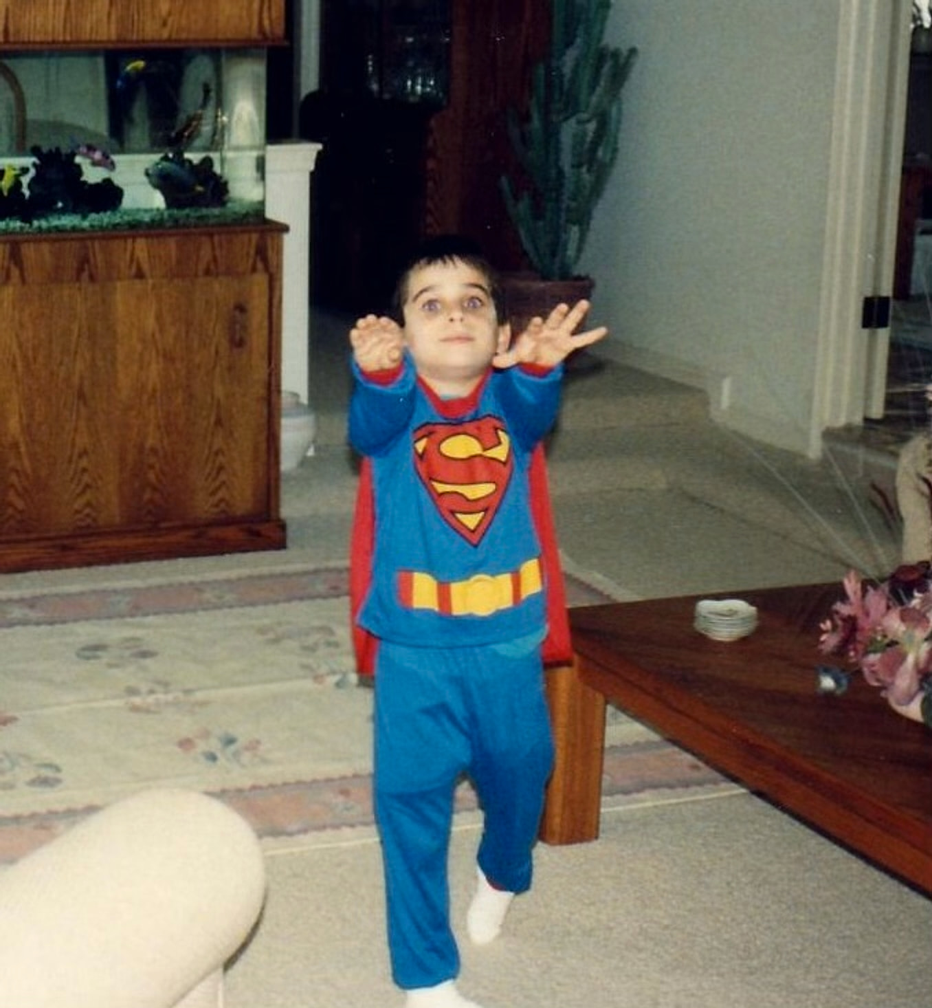 Ben Giroux as a little boy wearing his Superman shirt and small red cape with his arms outstretched